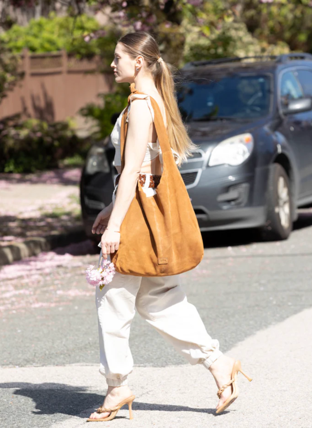 Woman holding flowers and wearing a stylish, large caramel-colored suede RISA upcycled handbag. The handcrafted shoulder bag complements her chic outfit, ideal for sustainable fashion lovers.