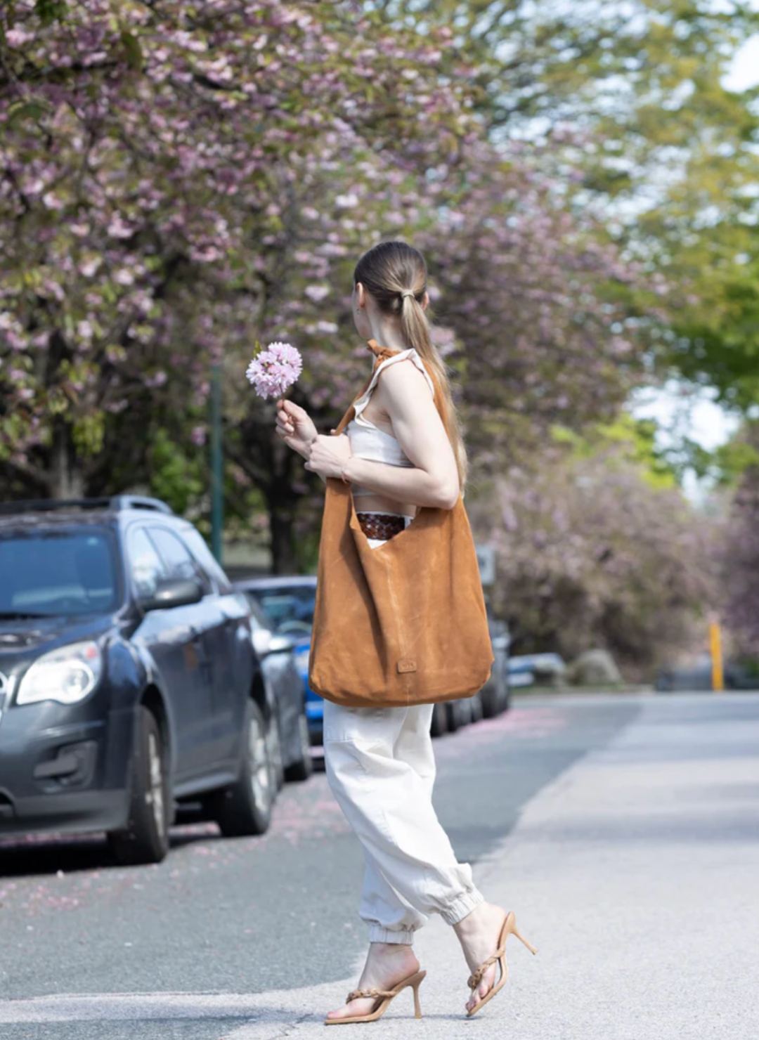 Woman holding flowers and wearing a stylish, large caramel-colored suede RISA upcycled handbag. The handcrafted shoulder bag complements her chic outfit, ideal for sustainable fashion lovers.