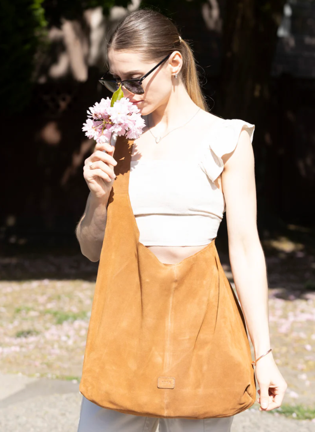 Woman holding flowers and wearing a stylish, large caramel-colored suede RISA upcycled handbag. The handcrafted shoulder bag complements her chic outfit, ideal for sustainable fashion lovers.