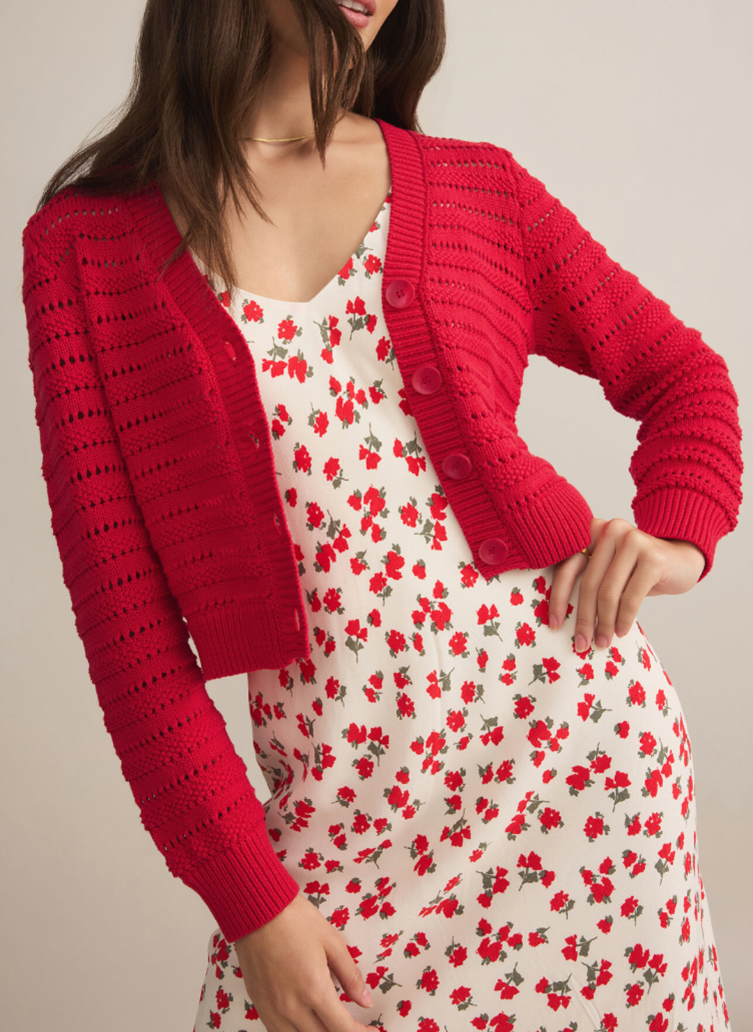 Model is wearing a red shorter length cardigan that buttons up in the front. Models is wearing the cardigan open over a flower printed white and red dress.