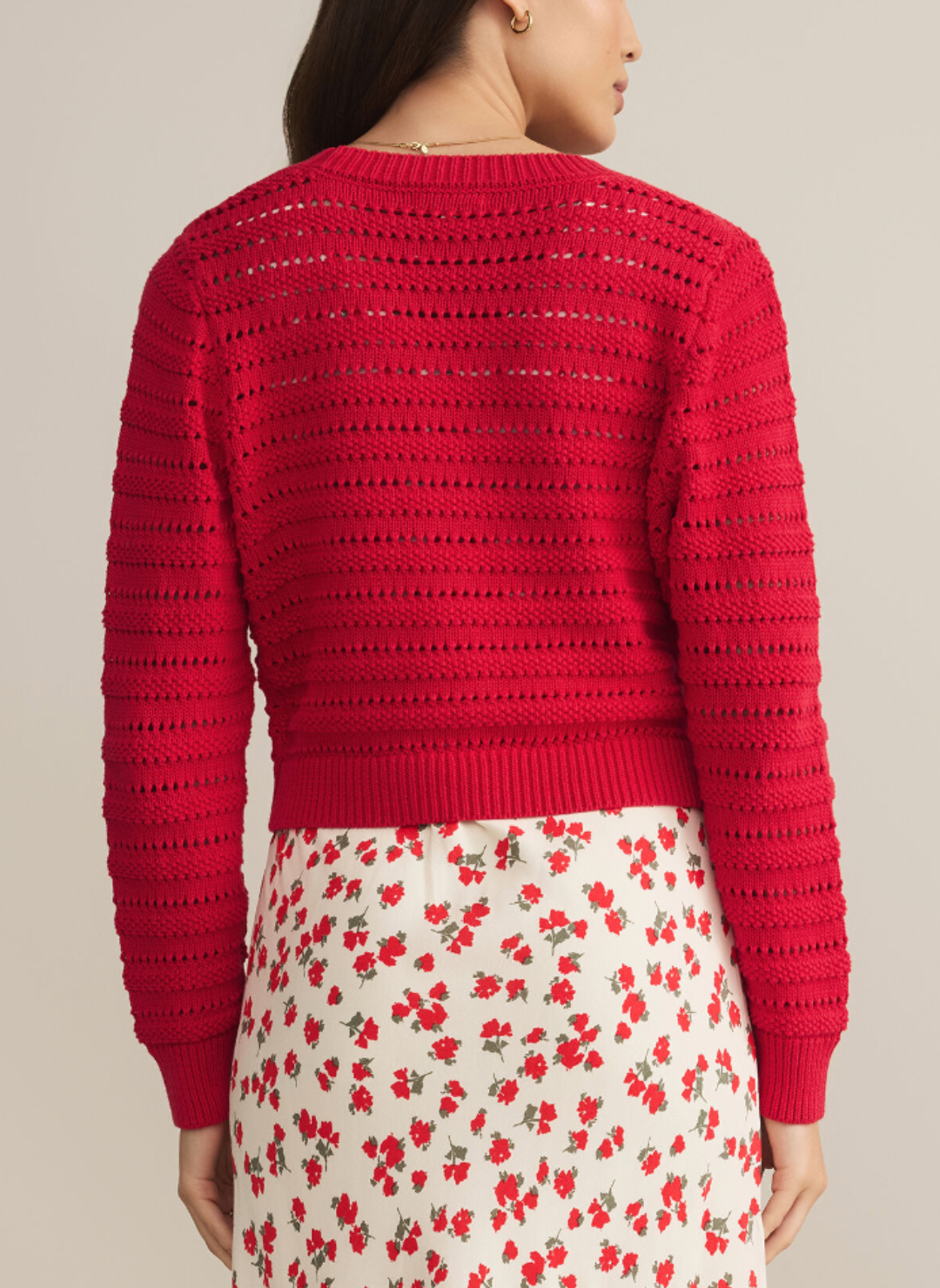 Model is wearing a red shorter length cardigan with detailed fabric throughout. Model is turned away showing the backside of the sweater.