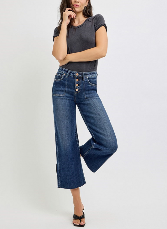 High rise cropped denim jeans with 4 buttons. The jeans are cropped and wide legged. Model is wearing a gray shirt either arm wrapped around her stomach. Models right leg is slightly bent behind her left leg.