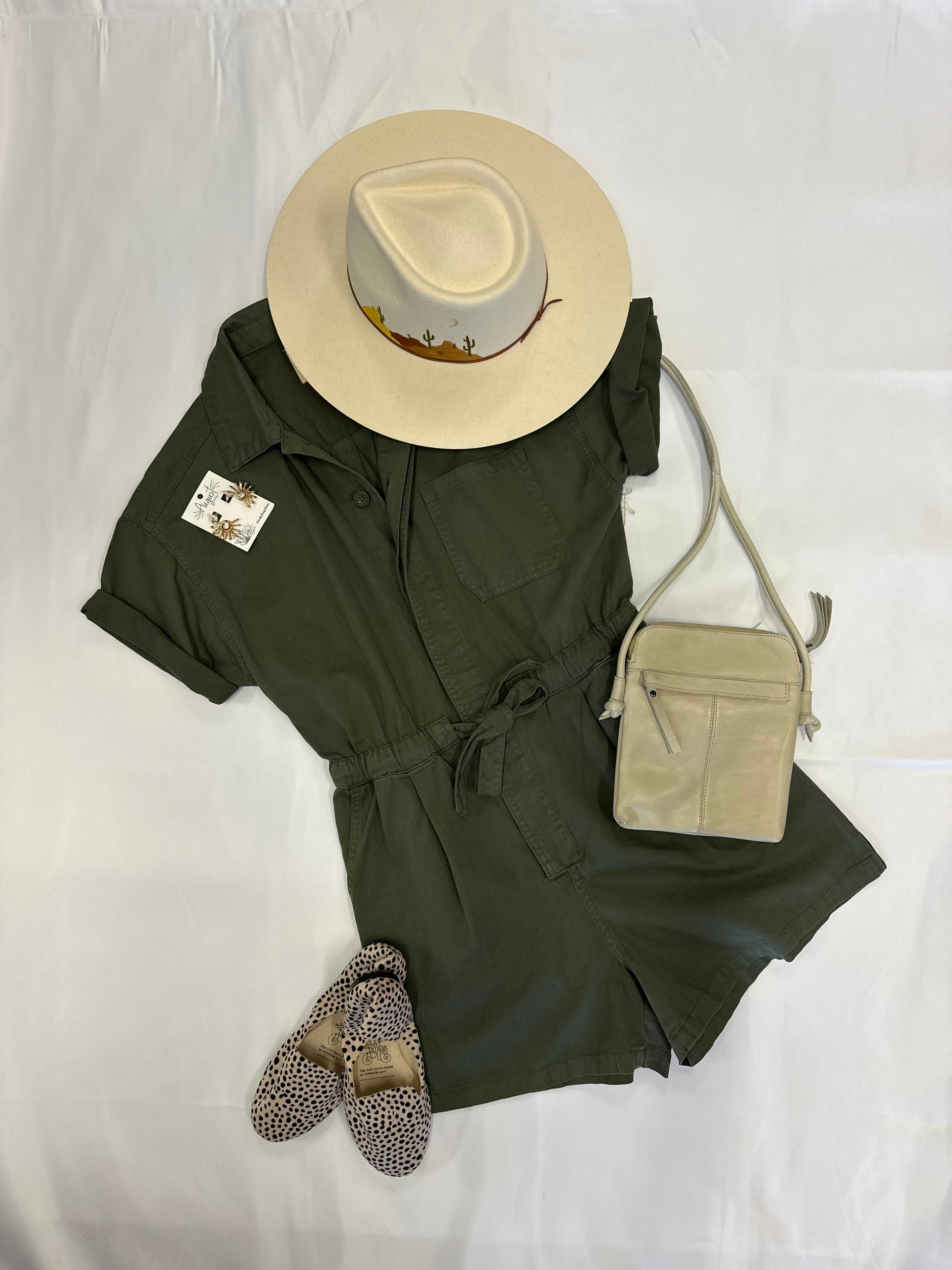 Front view of Army green romper accessorized with desert detailed hat, off white one strap bag, cheetah print flats, and gold earrings.   