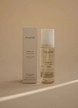 Mushie Baby Oil 145 mL bottle displayed beside its matching beige packaging on a neutral surface, showing the fragrance-free baby skincare product