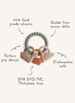 Mushie Dino Teething Ring product graphic showing the green textured silicone ring with three attached dinosaur charms in neutral earthy colors