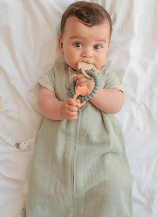 Baby holding and chewing the Mushie Dino Teething Ring, showing the flexible green silicone ring and three dinosaur-shaped charms during sensory play