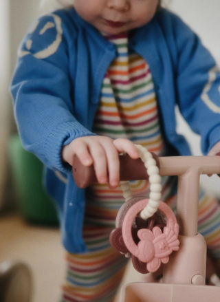 Child wearing a blue outfit and holding the Mushie Fairy Teething Ring, showing the textured silicone ring and pastel fairy-themed charms during play