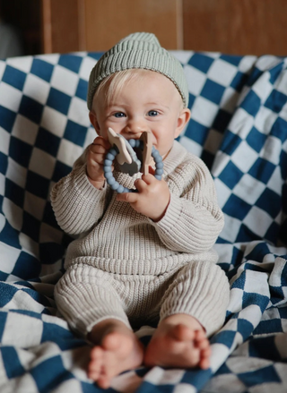 Baby holding the Mushie Space Teething Ring while sitting on a blue and white checkered blanket, showing the textured ring and space-themed charms