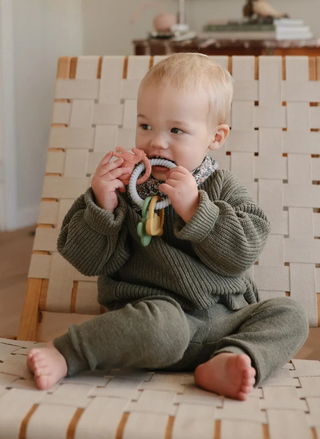 Baby holding and chewing a Mushie Western Teething Ring, showing the flexible textured ring and colorful Western-themed charms designed for teething and sensory exploration