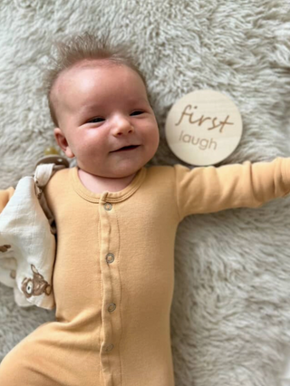 Baby in a yellow onesie lying on a fluffy surface with a 'first laugh' milestone plaque.