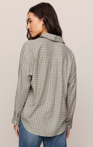 A woman shown from the back wearing an oversized long-sleeve button-up shirt with a small-scale green and cream gingham pattern, paired with blue jeans, against a plain light beige studio background.