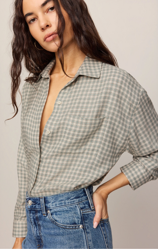 A woman wearing a muted sage green and cream gingham button-up shirt tucked into blue jeans, photographed against a plain light beige studio background.