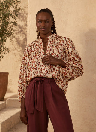 A woman wearing a Dress To cream blouse with a rust-red botanical pattern and dark burgundy high-waisted wrap pants, standing against a warm beige stucco wall.
