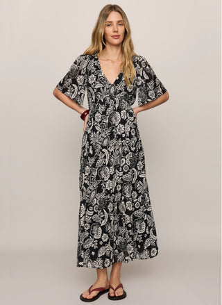 Front view of the Z Supply Lacey Floral Midi Dress in a black-and-ivory botanical print.
A woman modeling a black midi dress with a white botanical paisley pattern, featuring short flutter sleeves and a V-neckline, set against a plain light gray studio background.