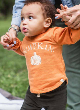 Child wearing an orange shirt with 'PUMPKIN' text and a pumpkin graphic, standing outdoors.