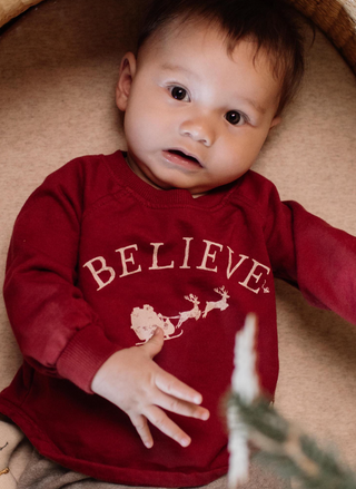Baby wearing Burt’s Bees Baby Believe Sweatshirt in red organic cotton with Santa sleigh and reindeer holiday graphic