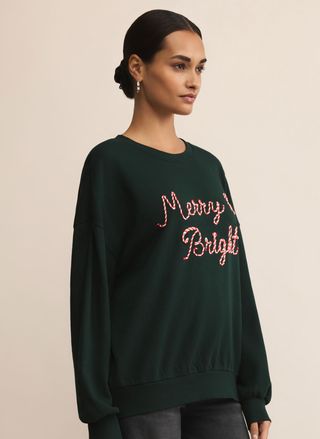 Woman wearing a dark green sweatshirt with 'Merry Bright' text in red on a beige background