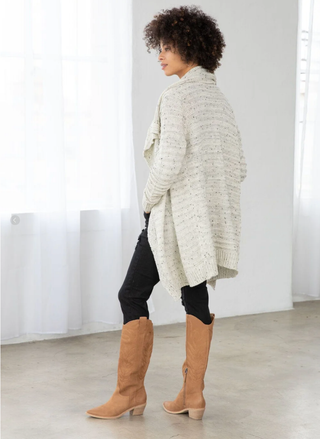 Person wearing a long cream cardigan, black pants, and tan knee-high boots in a minimalistic setting