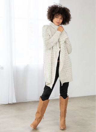 Woman wearing a long cream cardigan, black pants, and tan knee-high boots against a white background