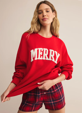 Woman wearing a red 'MERRY' sweatshirt and plaid shorts on a beige background