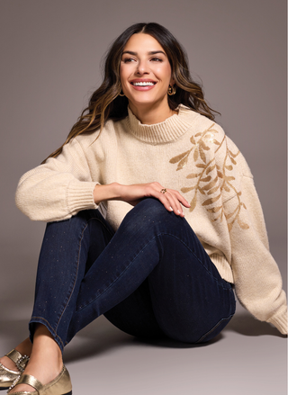 Woman wearing a beige sweater with floral patterns and blue jeans, sitting on the floor against a gray background.
