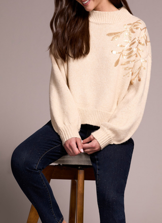 Woman wearing a beige sweater with floral patterns sitting on a stool against a plain background