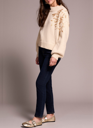 Woman wearing a beige sweater and dark jeans against a plain background