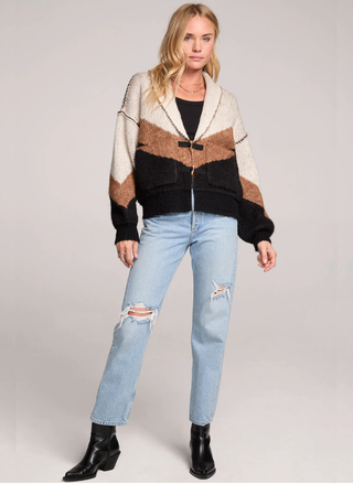 Woman wearing a patterned cardigan, black top, light blue jeans, and black boots on a white background