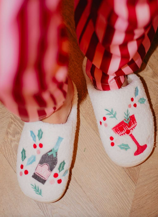 White slippers with festive designs worn by a person in red plaid pants on a wooden floor.