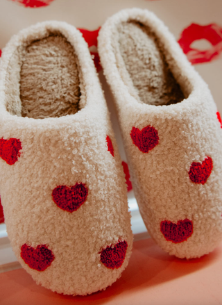 Pair of beige slippers with red heart patterns on a light background. Valentine heart slippers soft plush indoor shoes.