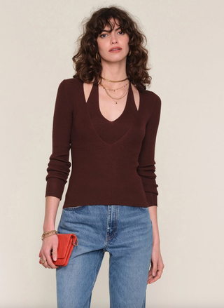 Woman wearing a brown sweater and blue jeans, holding a red clutch against a plain background