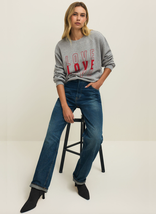 Woman wearing a gray sweatshirt with 'LOVE' text and blue jeans, sitting on a stool against a white background.