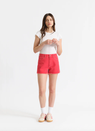 Model wearing Daze Denim Trouble Maker high-rise shorts in Chili Pepper red with a white ruffle tee and sneakers at August Boutique.
