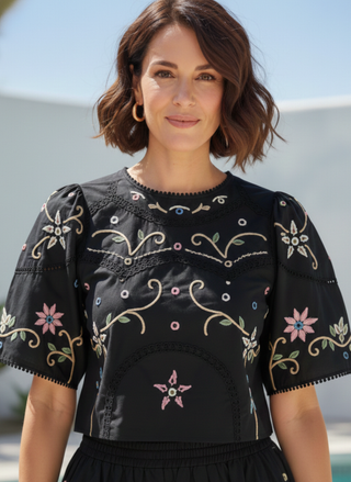 Adult woman in a desert setting wearing a black short-sleeved top with colorful floral embroidery and matching midi skirt.  Woman wearing a black top with colorful embroidery against a clear sky.
