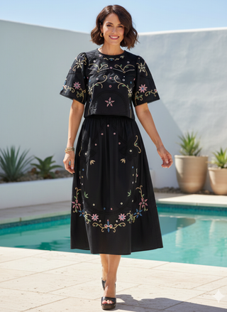 Adult woman in a desert setting wearing a black short-sleeved top with colorful floral embroidery and matching midi skirt by a pool.