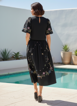 Woman wearing a black dress with floral embroidery by a poolside.