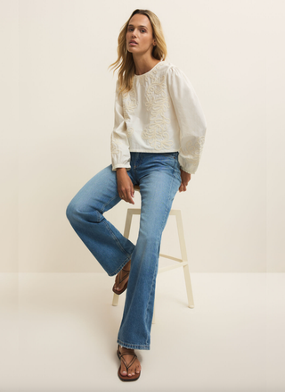 Woman wearing a white blouse and blue jeans sitting on a stool against a plain background