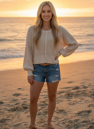 Woman standing on a beach at sunset wearing a beige cardigan and denim shorts.