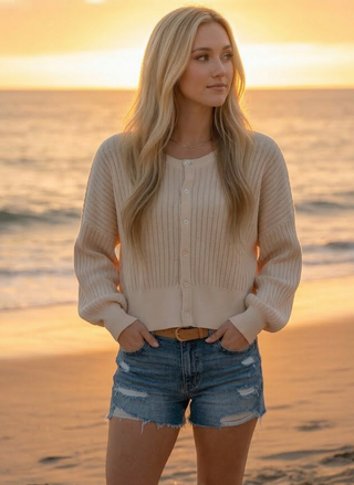 Woman wearing the Weslynn Collective Sara cream organic cotton cardigan with balloon sleeves and button front standing on a beach at sunset.