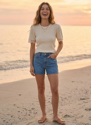 Woman standing on a beach at sunset wearing a white top and blue shorts.