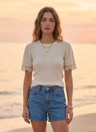 Woman standing on a beach wearing Weslynn Giselle Sweater Tee in beige and denim shorts with a sunset in the background at the beach