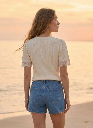 Woman standing on a beach at sunset wearing a beige top and blue shorts.