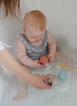 A lifestyle shot of a baby in a bubble bath playing with colorful silicone space toys including a peach planet and grey astronaut.