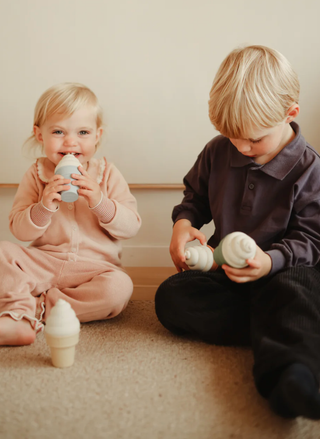 Two young children sitting on a rug playing with Mushie silicone ice cream toys in muted pastel colors.