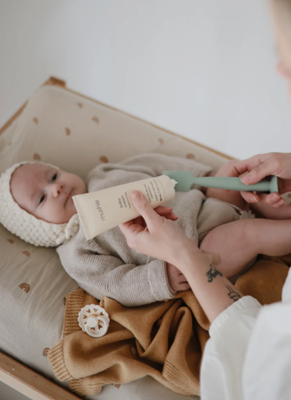 Mushie Baby Cream sitting next to a wooden brush and a soft swaddle on a neutral-toned changing table.