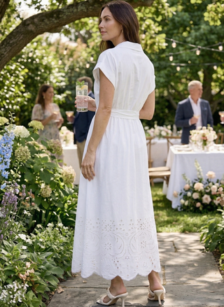 Woman in a white dress standing outdoors in a garden setting with tables and people in the background.