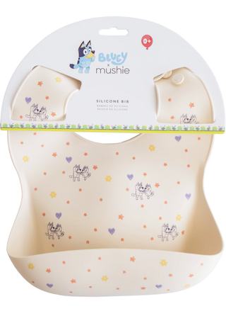 A cream-colored Mushie x Bluey silicone baby bib featuring Bluey and Bingo characters, stars, and hearts with a paper marketing hanger.