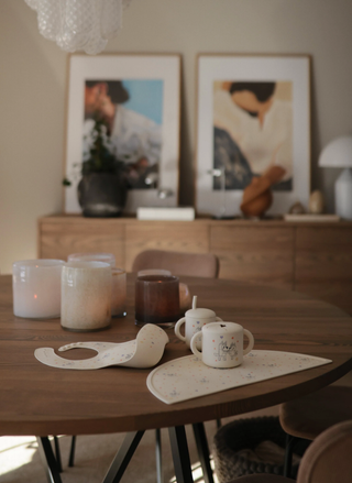 Dining table with mugs, a bib, and placemats in a room with framed pictures on the wall.