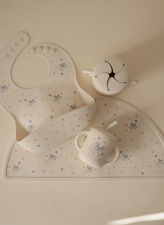 A cream-colored Mushie x Bluey silicone baby bib featuring Bluey and Bingo characters, stars, and hearts, displayed alongside a matching mealtime set including a sippy cup, snack cup, and placemat.