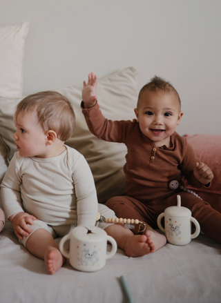 Two babies sitting on a bed with toys and sippy cups.